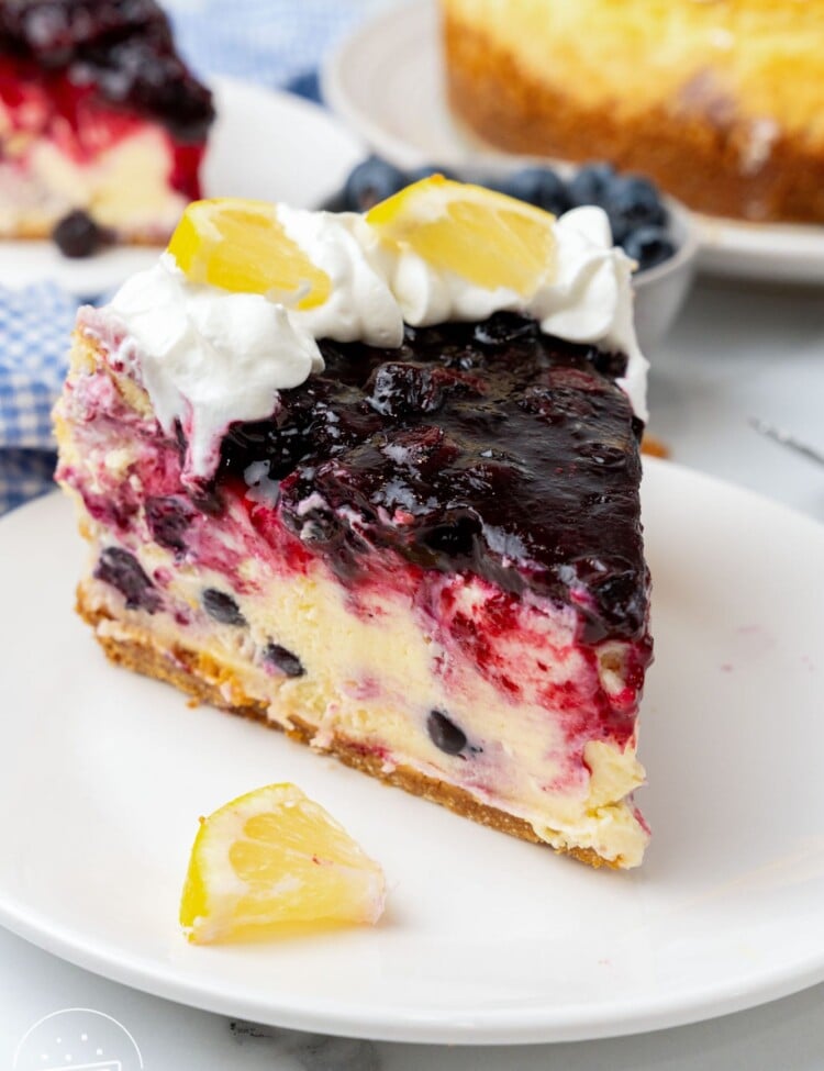 one slice of blueberry lemon cheesecake on a plate. The edge has whipped cream and fresh lemon wedges.