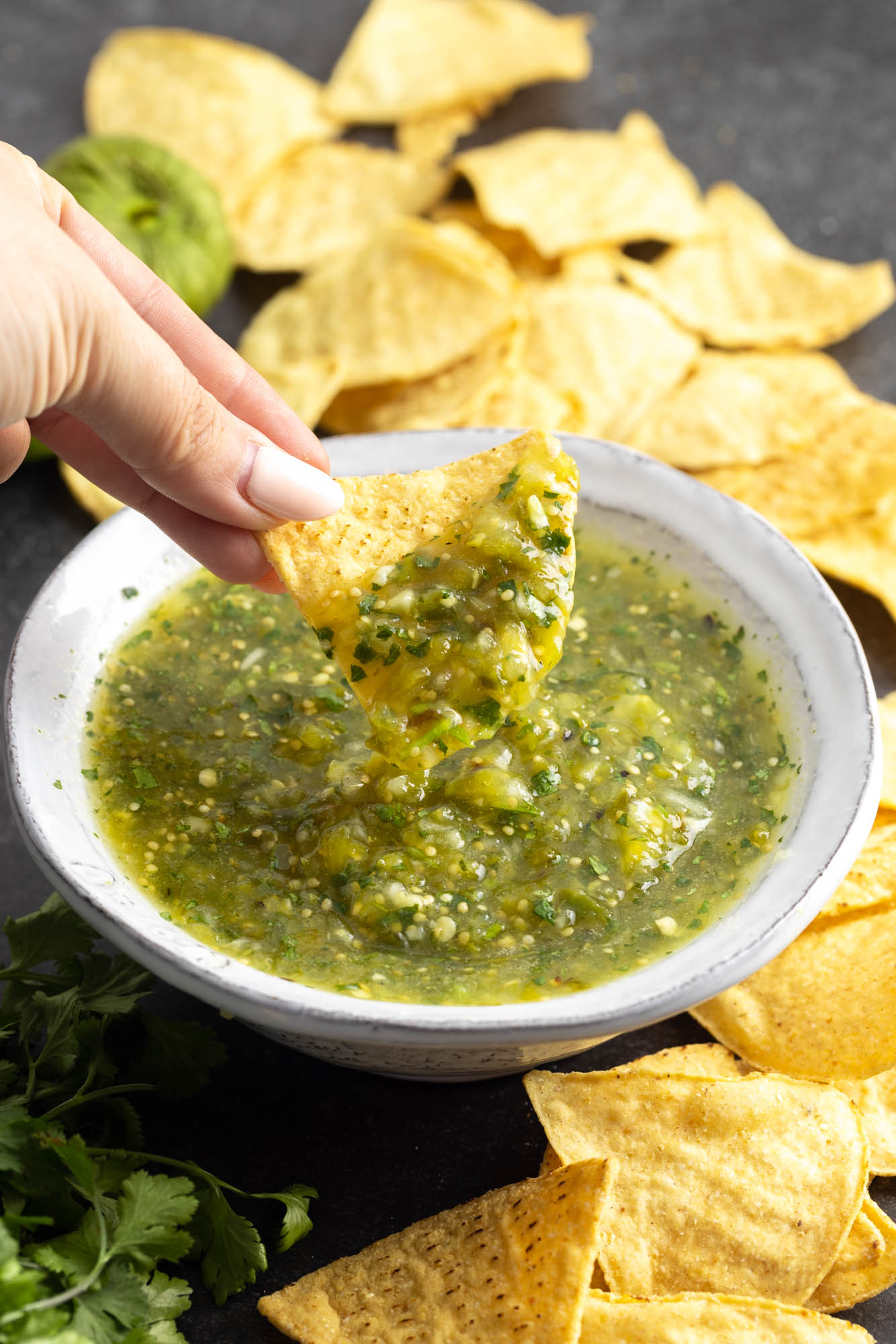 Dipping a tortilla chip in a bowl filled with mexican salsa verde, with more chips on the sides of the bowl.
