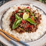 a plate of rice topped with pad kra pao, garnished with thai basil, sesame seeds, and lime wedges. Blue patterned chopsticks are on the side of the plate.