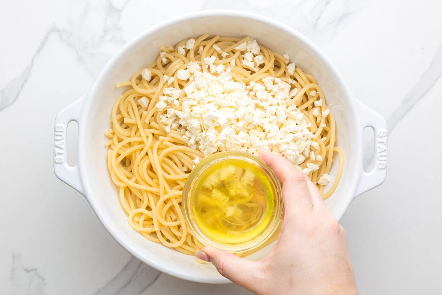 Feta and oil added to cooked Pastitsio pasta in a white mixing bowl.