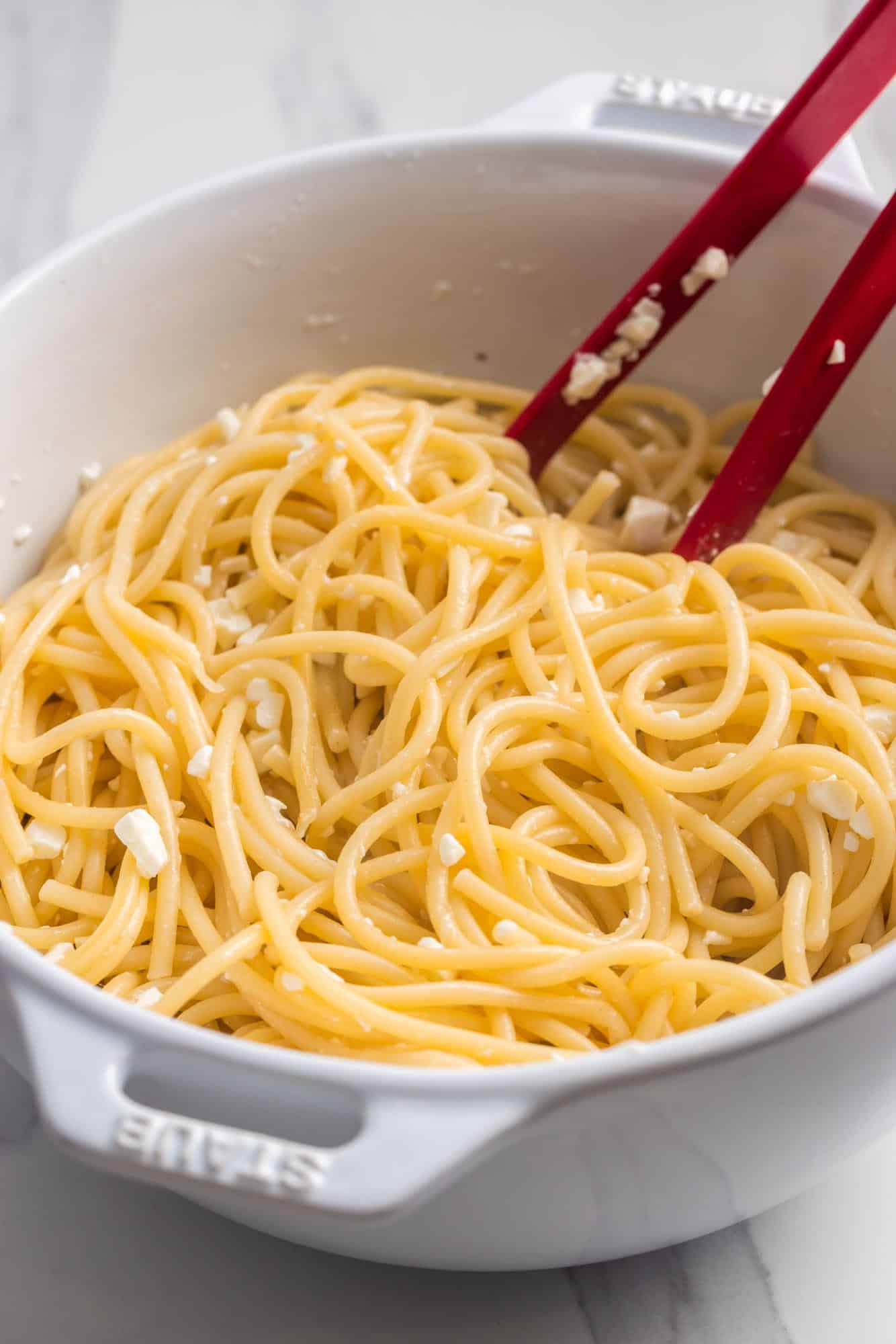 long pasta tossed with feta cheese and oil in a white mixing bowl