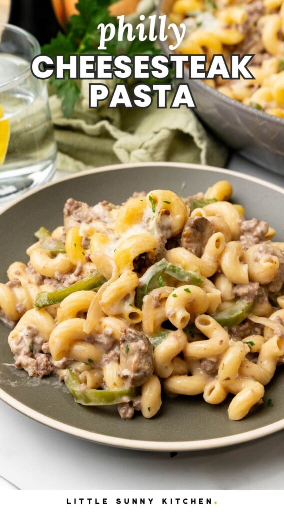 a serving of cheese steak pasta with peppers and onions on a dark ceramic plate. Text overlay says "philly cheesesteak pasta"