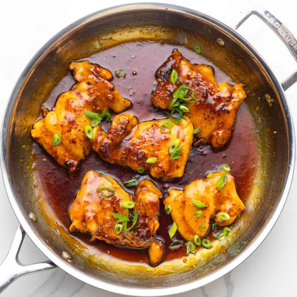 Boneless skinless chicken thighs cooking in a large stainless steel skillet in a apricot glaze, garnished with sliced green onion.