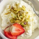 Overhead shot of Ashta cream served in a white bowl with a dessert spoon, garnished with crushed pistachios and 2 slices of fresh strawberry