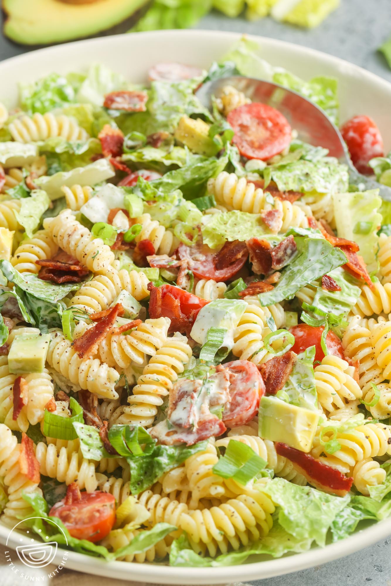 a large white serving bowl of creamy homemade BLT pasta salad with lettuce, cherry tomatoes, avocado, bacon, and rotini pasta,