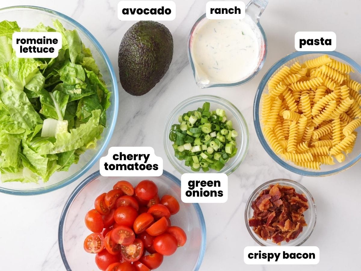 The ingredients needed to make BLT pasta salad, including pasta, ranch dressing, avocado, baking, lettuce, tomato, and green onions. All ingredients are in separate bowls.