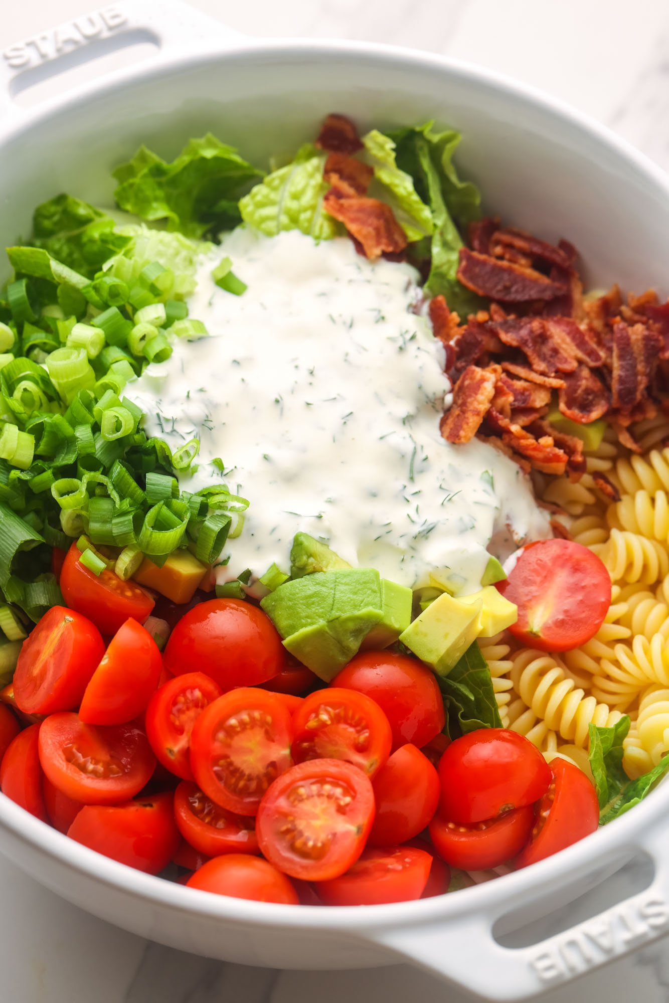a white ceramic bowl holding cooked pasta, cooked bacon, chopped tomato, green onion, lettuce, and avocado. Ranch dressing is poured into the middle.
