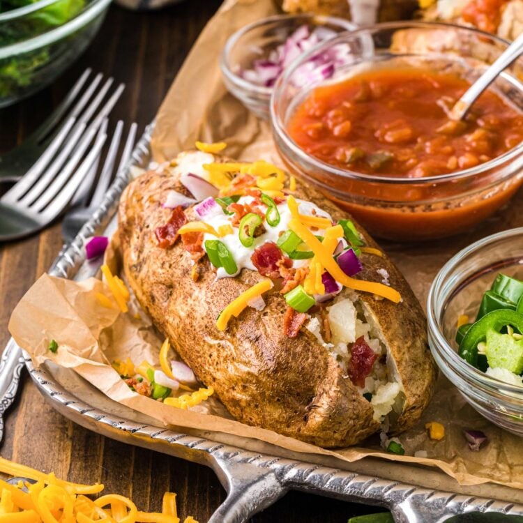 a silver tray lined with brown parchment paper holding a loaded baked potato, and small bowls of toppings including salsa, jalapenos, red onions.