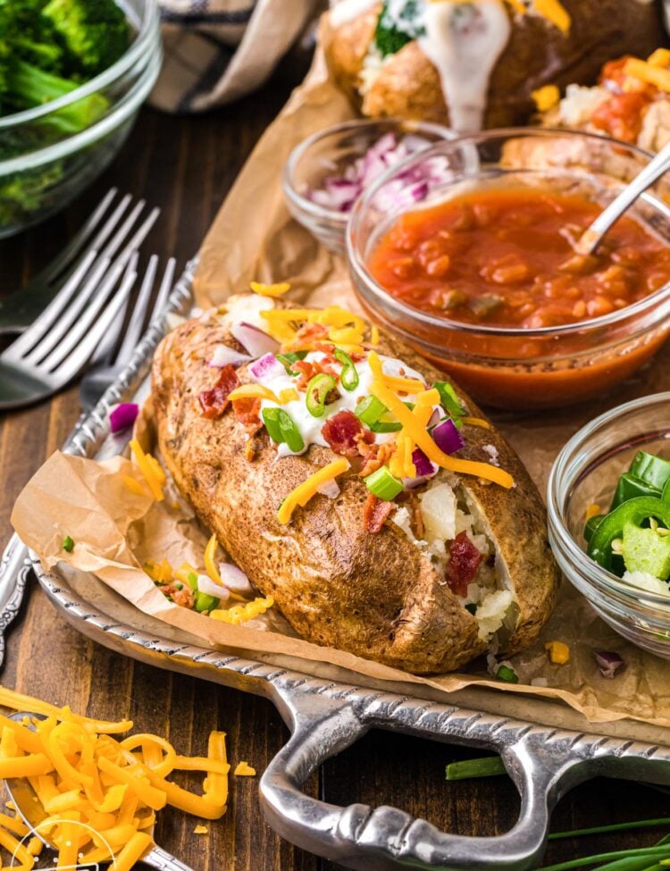 a silver tray lined with brown parchment paper holding a loaded baked potato, and small bowls of toppings including salsa, jalapenos, red onions.
