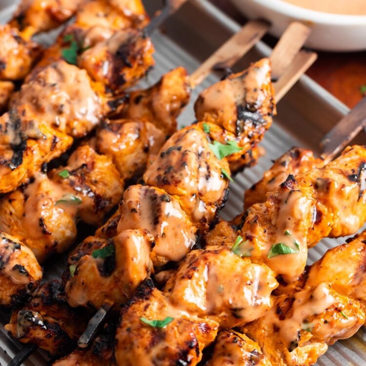 a ridged metal baking pan of grilled chicken skewers with bang bang sauce, garnished with chopped parsley.