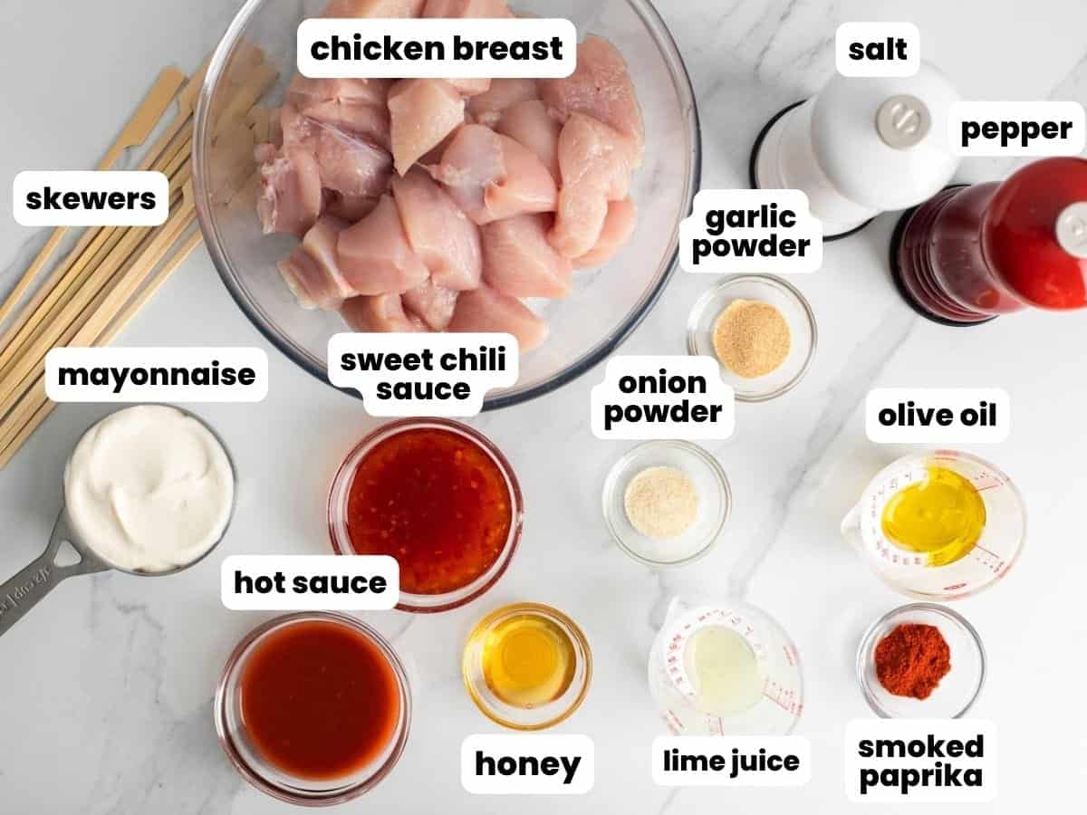 the ingredients needed to make bang bang chicken skewers, all measured into small bowls and arranged on a marble counter, along with wooden skewers.