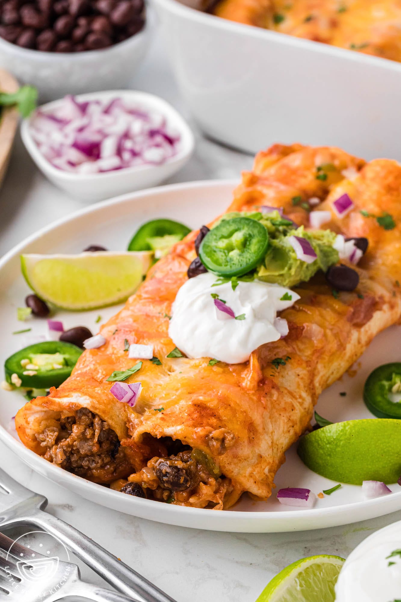 a plate holding two cheesy beef enchiladas topped with beans, sour cream, guacamole, and a sliced jalapeno.