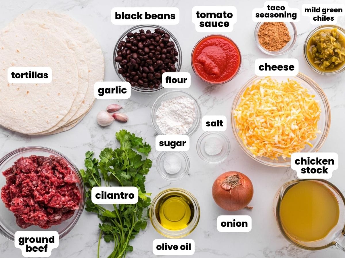The ingredients needed to make beef enchiladas arranged on a counter, including ground beef, flour tortillas, shredded cheese, black beans, and seasonings.