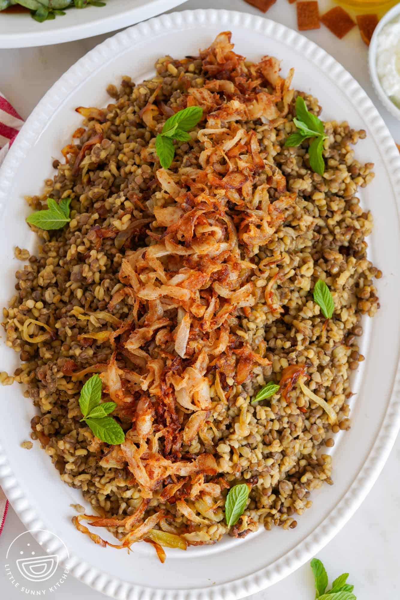 Overhead shot of Bulgur Mujadara served in a large oval white plate, topped with crispy onions and fresh mint leaves as a garnish