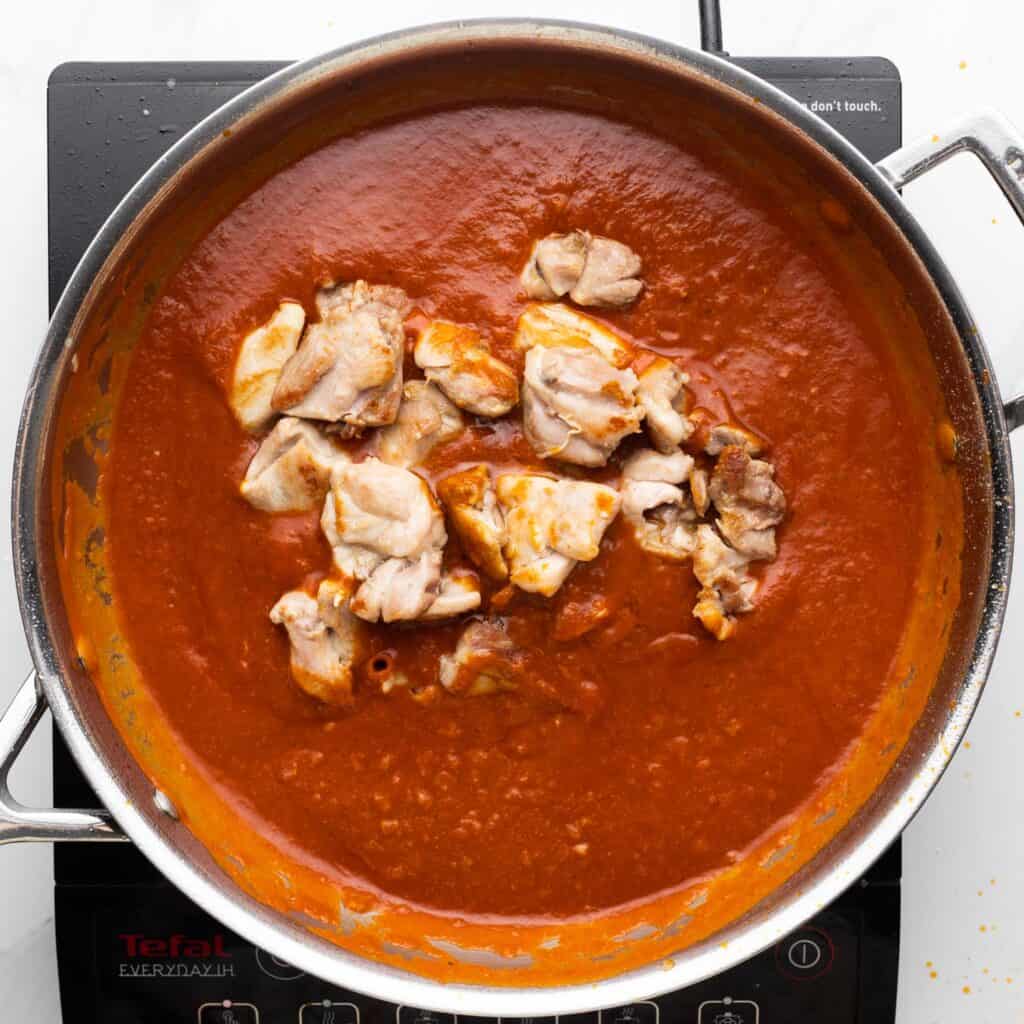 cooked chicken added to a deep red butter chicken sauce in a skillet set over an electric burner