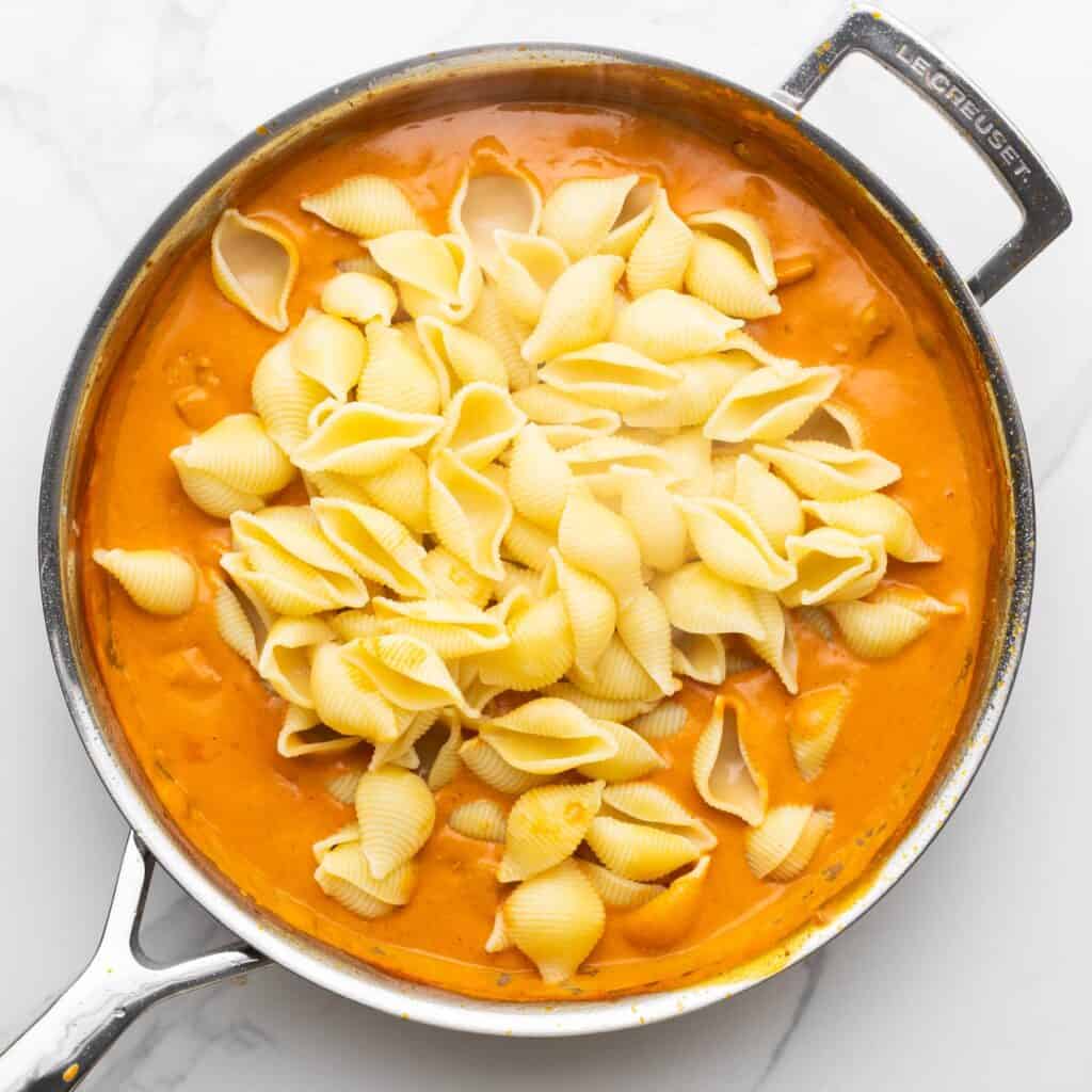 cooked shell pasta added to creamy butter chicken sauce in a silver skillet.