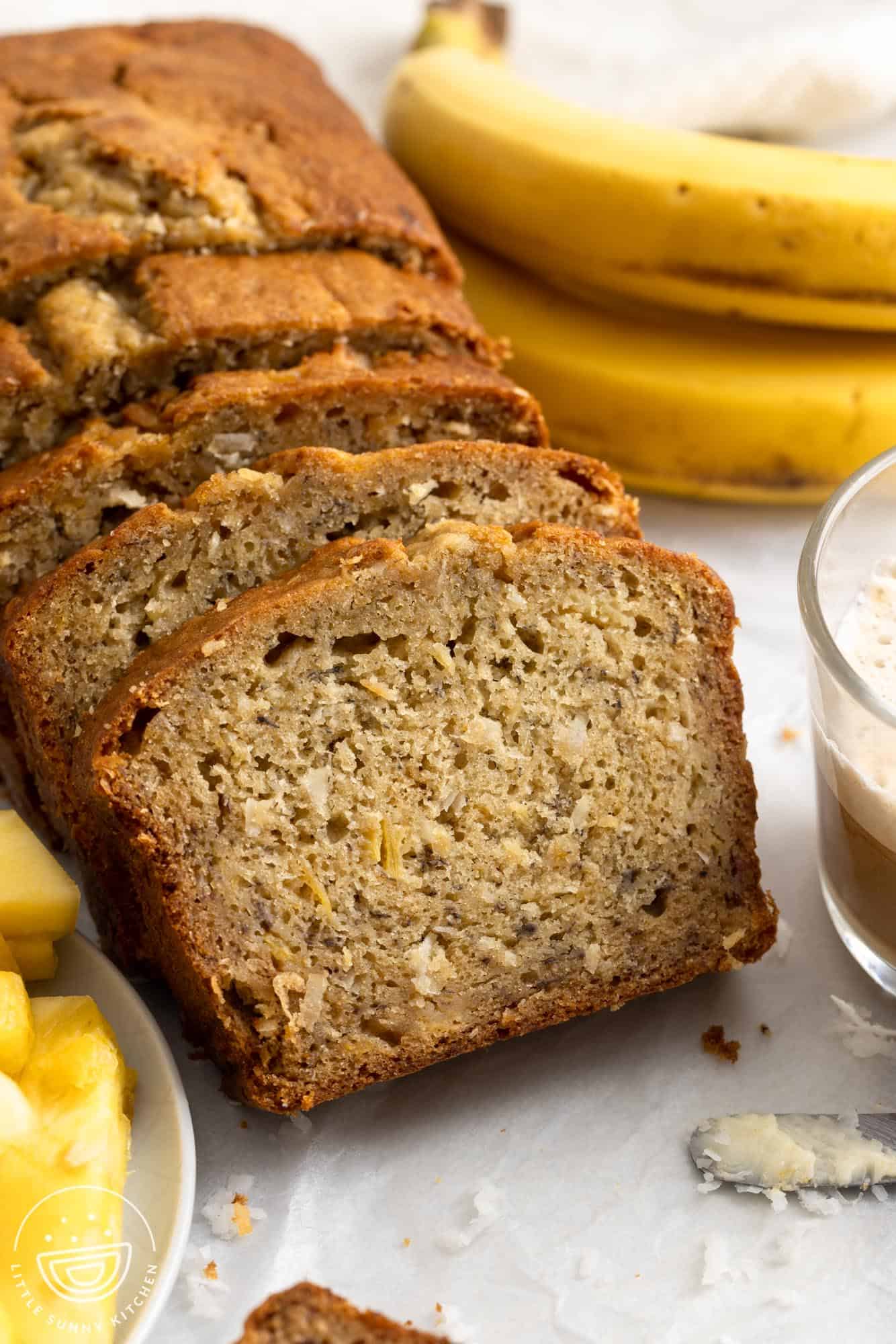 a loaf of hawaiian banana bread with pineapple and coconut, sliced, next to fresh bananas.