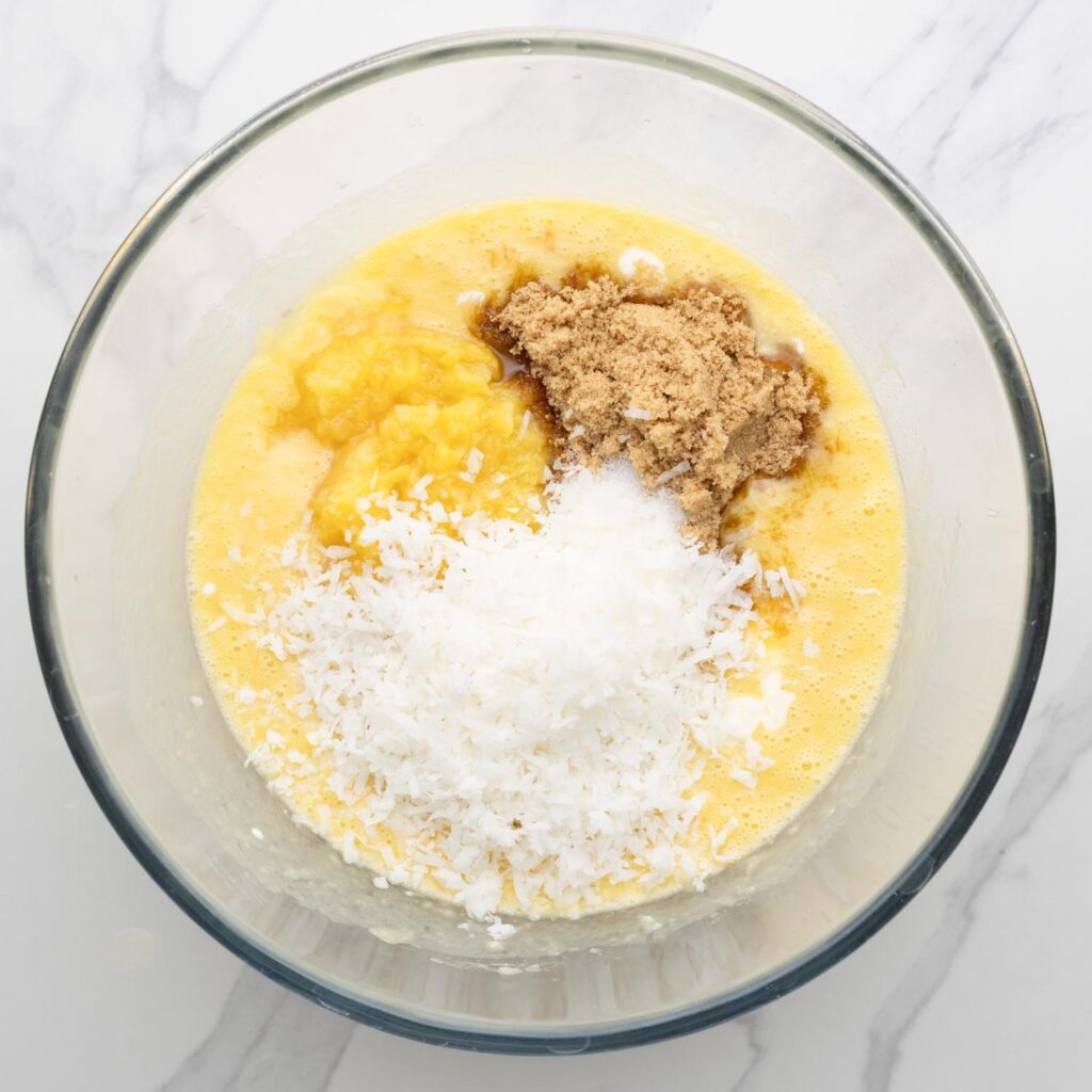 a glass bowl holding ingredients for hawaiian banana bread batter, including shredded coconut, crushed pineapple, and brown sugar.