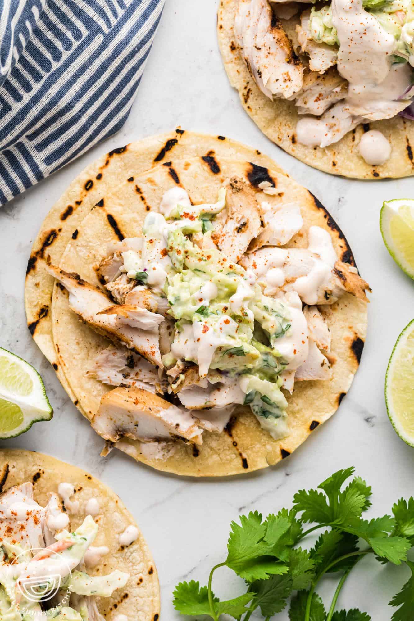 grilled corn tortillas topped with flaked mahi mahi, slaw, and creamy sauce. Fresh cilantro and lime wedges are next to the tacos on a marble counter.