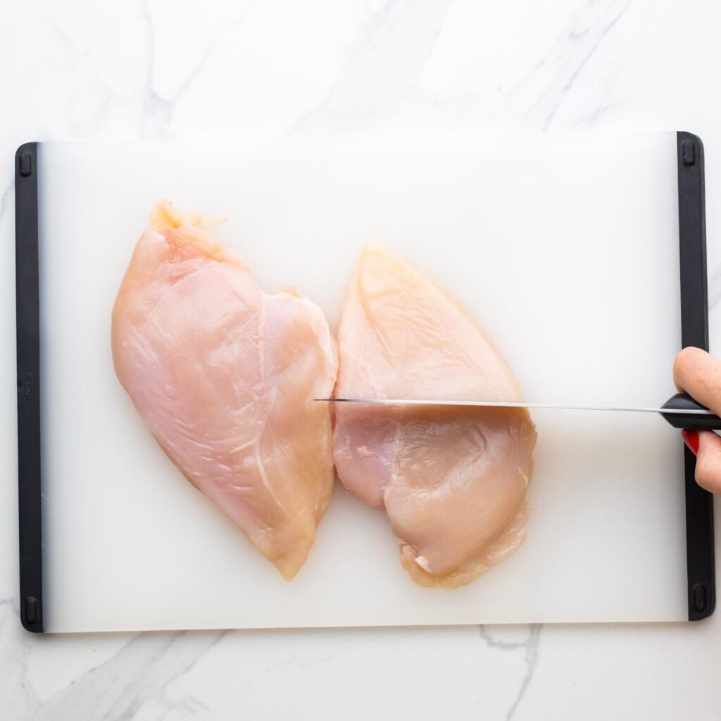 2 chicken breasts on a cutting board, and a knife cutting one in half