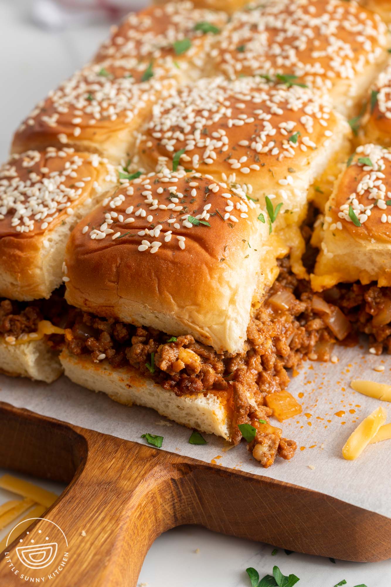 A wooden cutting board with a sheet of parchment holding rows of baked Sloppy Joe Sliders made with Hawaiian rolls.