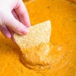 a hand dipping a tortilla chip into a crock pot filled with queso dip.