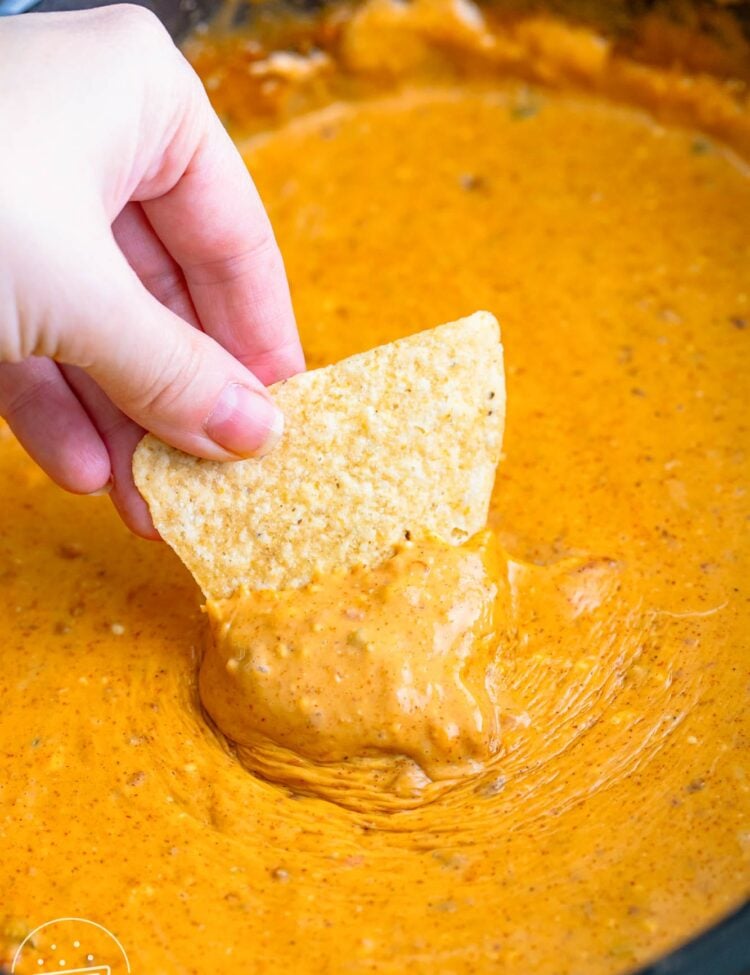 a hand dipping a tortilla chip into a crock pot filled with queso dip.
