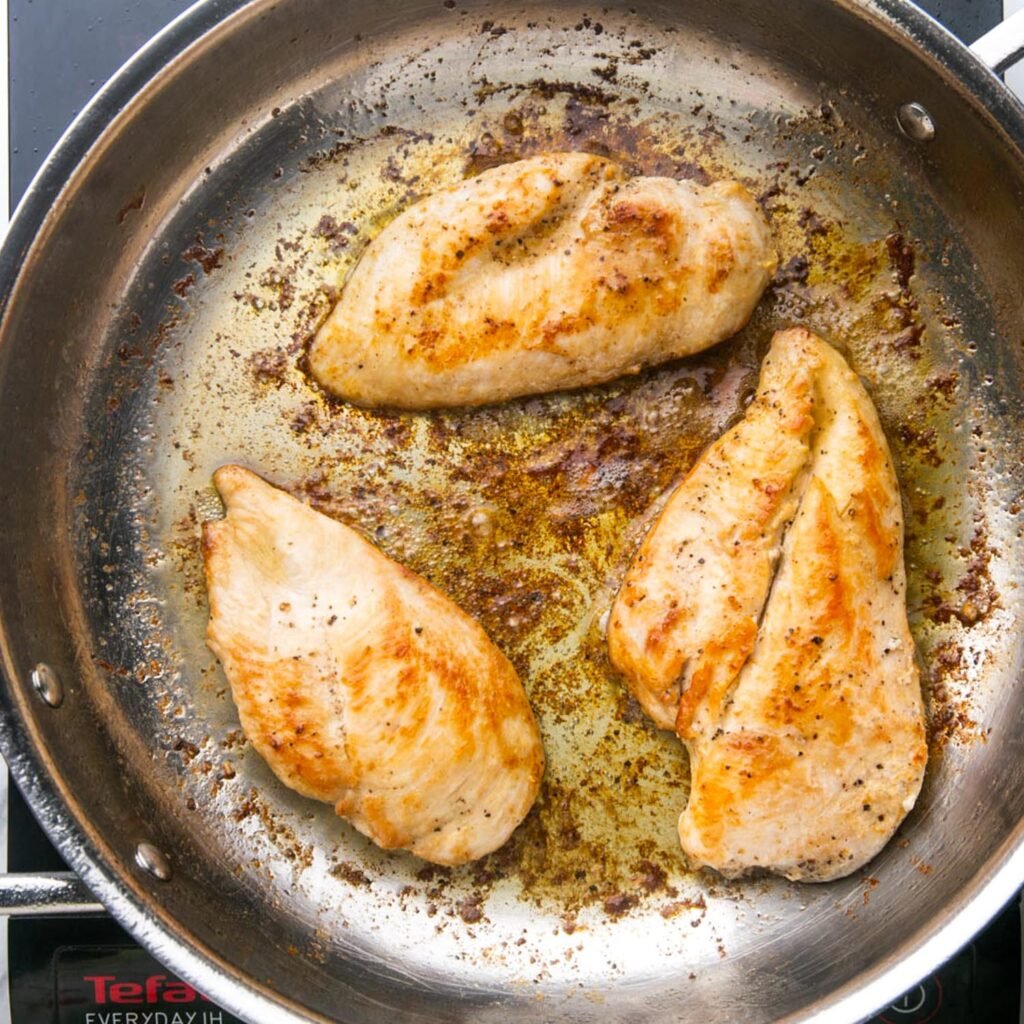 three chicken cutlets cooking in a steel skillet with oil and butter.