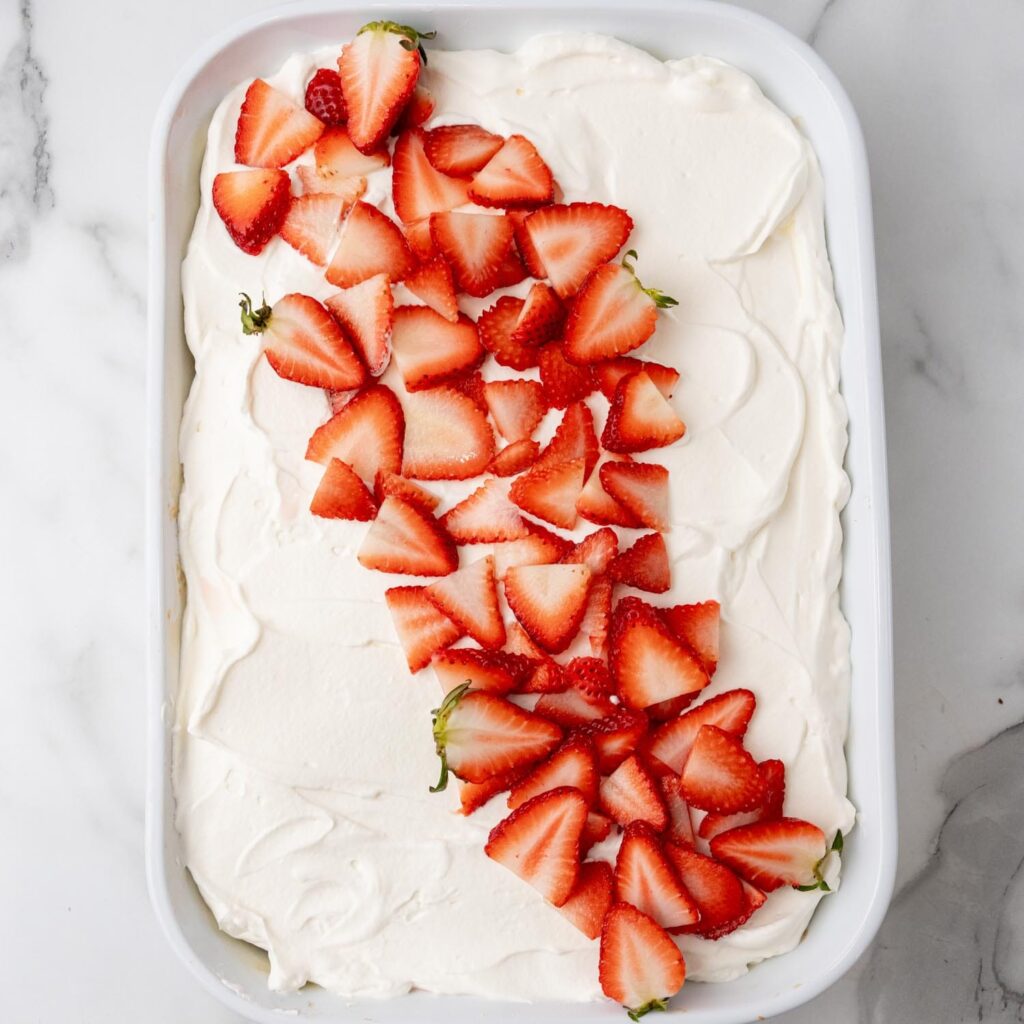a cake in a white baking dish, covered with whipped cream frosting and a diagonal line of sliced strawberries for decoration. 