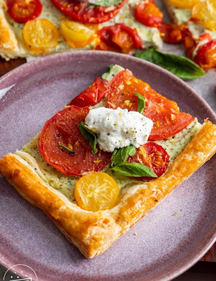 a square piece of tomato tart on a purple serving plate. The tart is topped with a dollop of ricotta, basil leaves, salt and pepper.