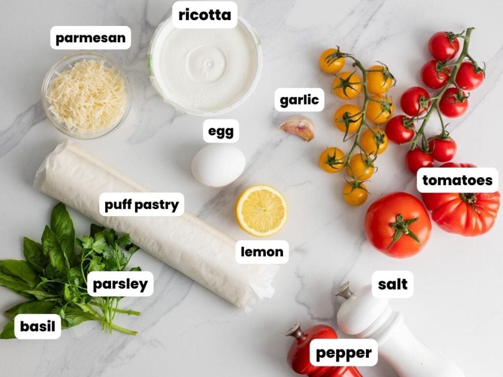 Fresh heirloom and cherry tomatoes along with all of the ingredients needed to make a homemade puff pastry tomato tart, arranged on a marble counter