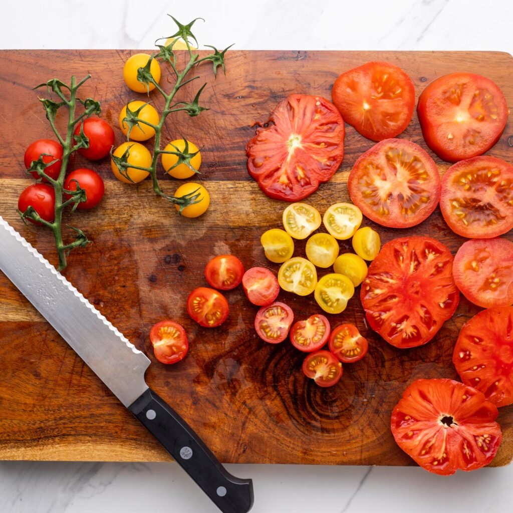 a wooden cutting board with a serrated knife and sliced tomatoes of various sizes and colors.