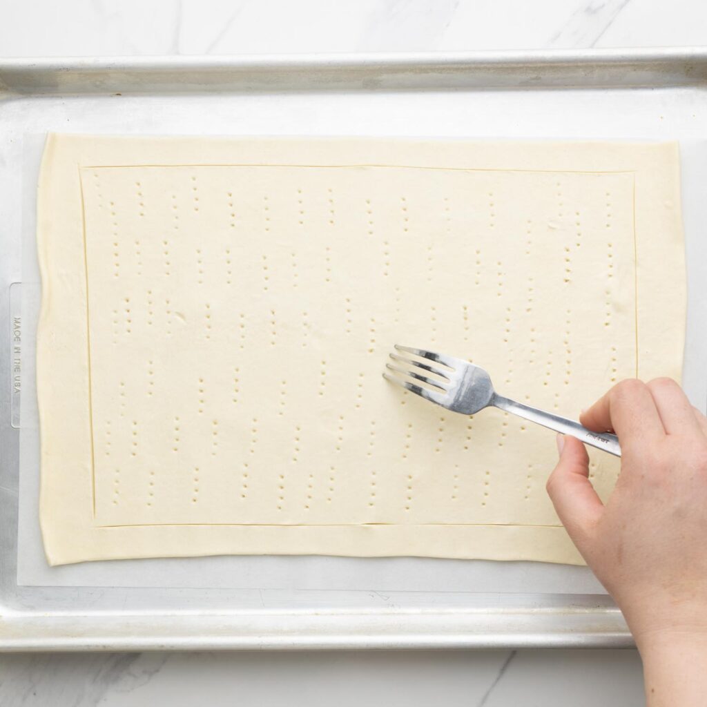 a roll of puff pastry on a parchment lined baking sheet. It has been scored around the sides to create a curst and a fork is being used to poke holes in the center.