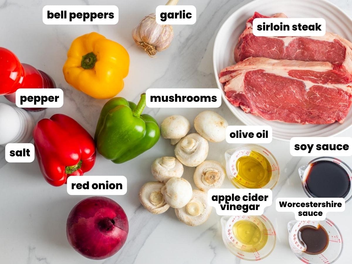 a plate of raw sirloin steak with fresh mushrooms, bell peppers, and a red onion. Ingredients for a marinade for beef kabobs are measured into small measuring cups. Ingredients are arranged on a marble counter, viewed from above.