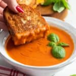a sourdough grilled cheese sandwich dipped into a ceramic bowl of tomato soup that is garnished with basil leaves.