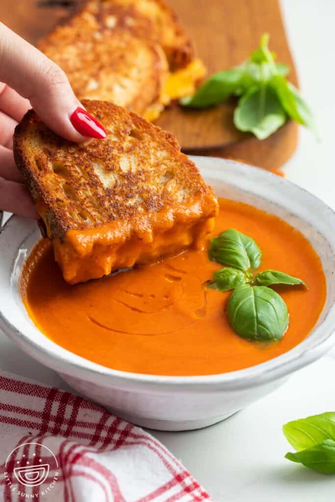 a sourdough grilled cheese sandwich dipped into a ceramic bowl of tomato soup that is garnished with basil leaves.