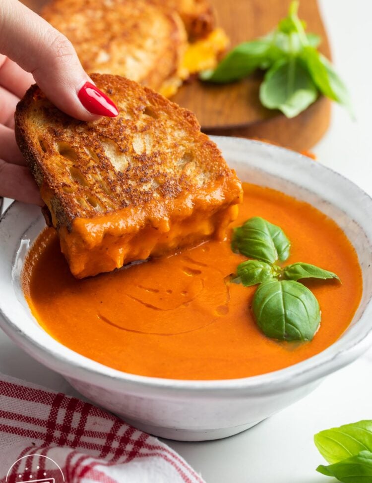 a sourdough grilled cheese sandwich dipped into a ceramic bowl of tomato soup that is garnished with basil leaves.