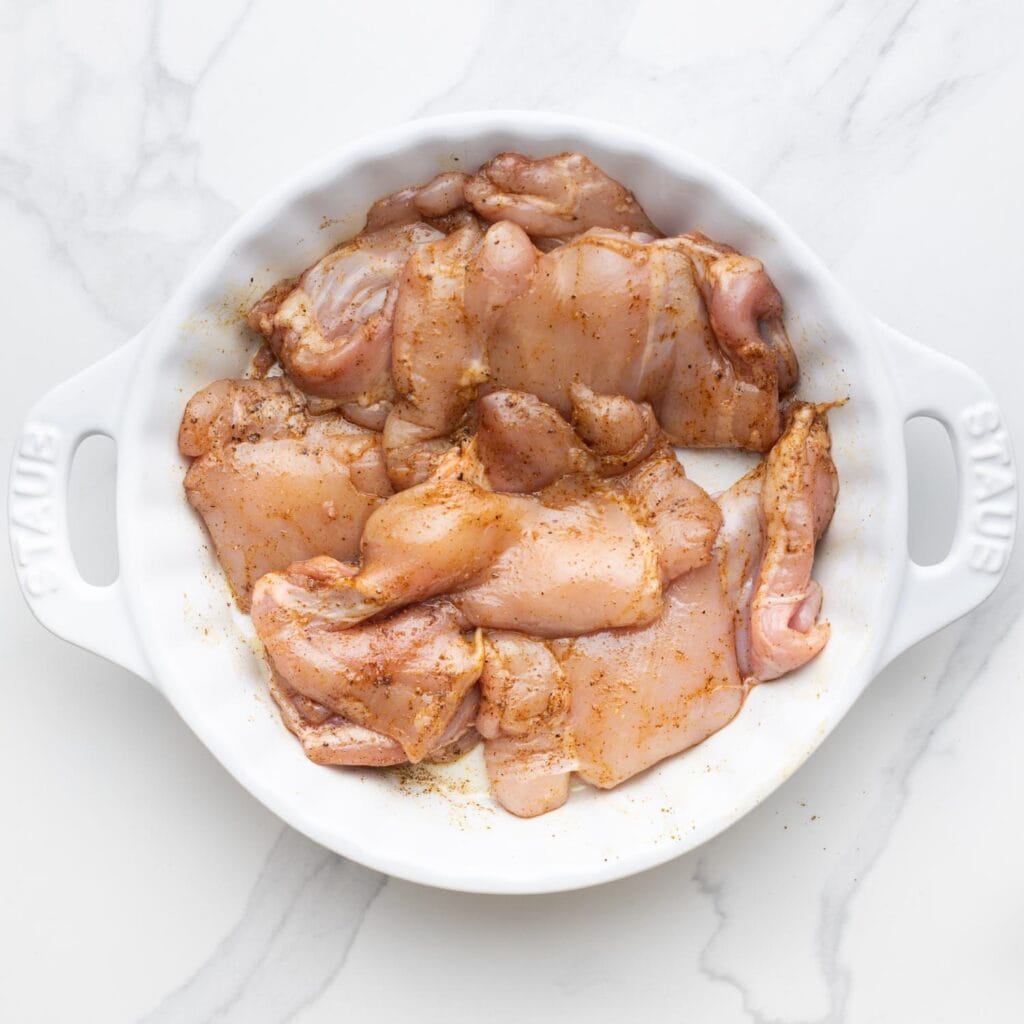 Boneless skinless chicken thighs, raw and seasoned in a white pie plate.