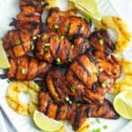 a large white platter of grilled huli huli chicken with grilled pineapple rings and lime wedges around the sides of the plate.