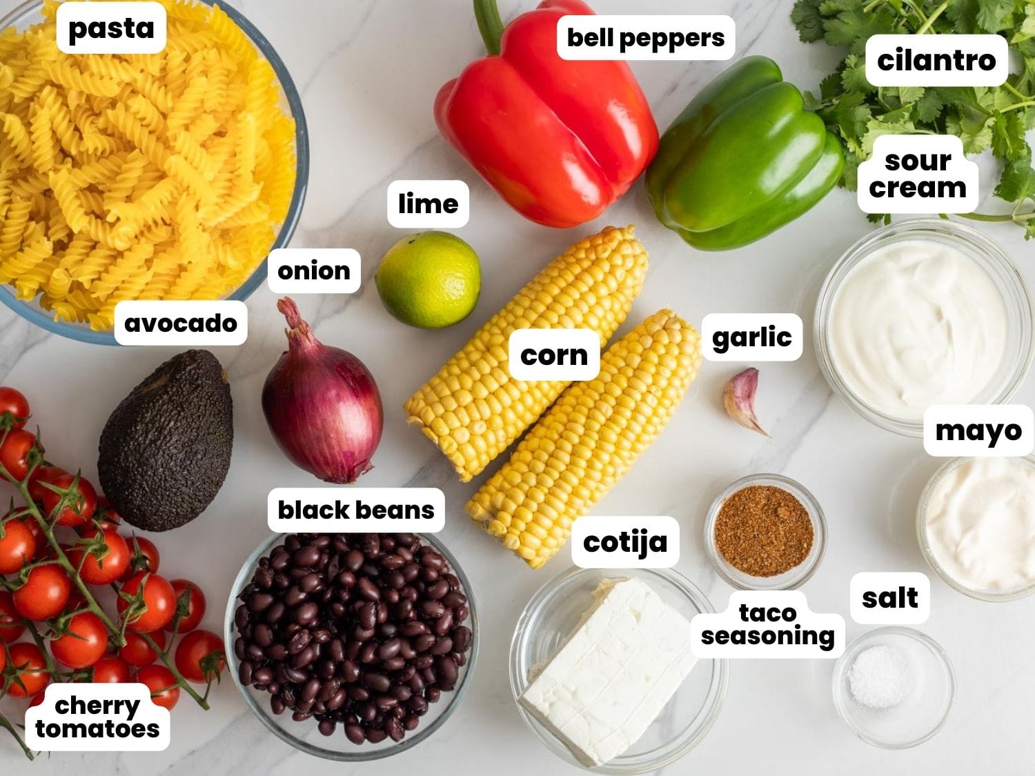 The ingredients needed to make mexican pasta salad including tomatoes, peppers, corn, and beans, are arranged on a marble counter top, viewed from above.