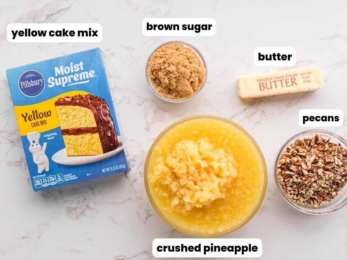 The five ingredients needed to make pineapple dump cake: Yellow cake mix, crushed pineapple, brown sugar, pecans, and butter, in separate bowls, on a marble counter.