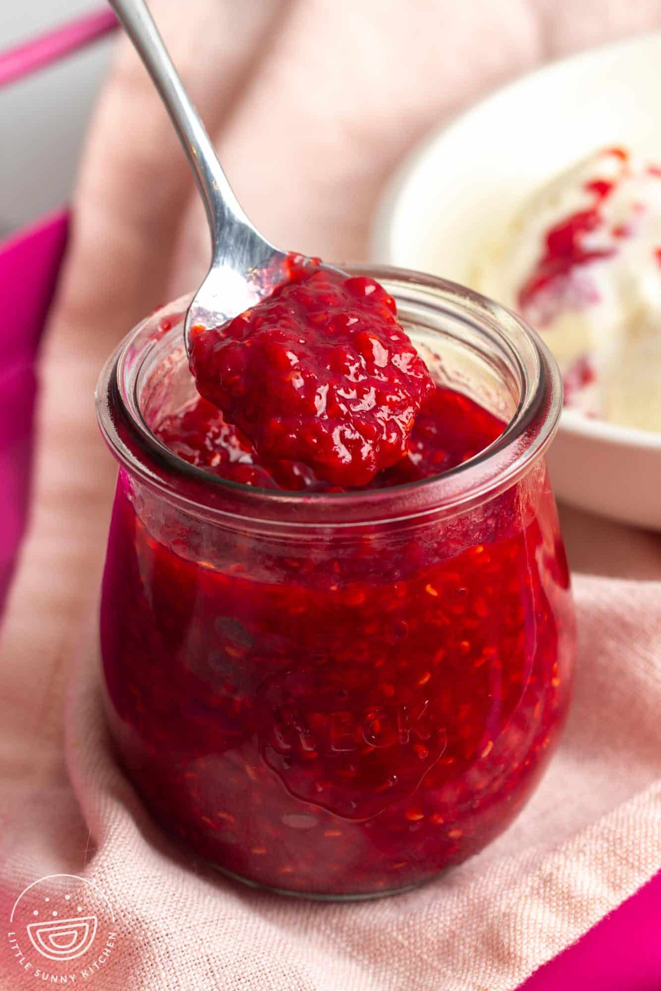 a small rounded weck jar of raspberry compote. A spoon is lifting out some of the sauce.