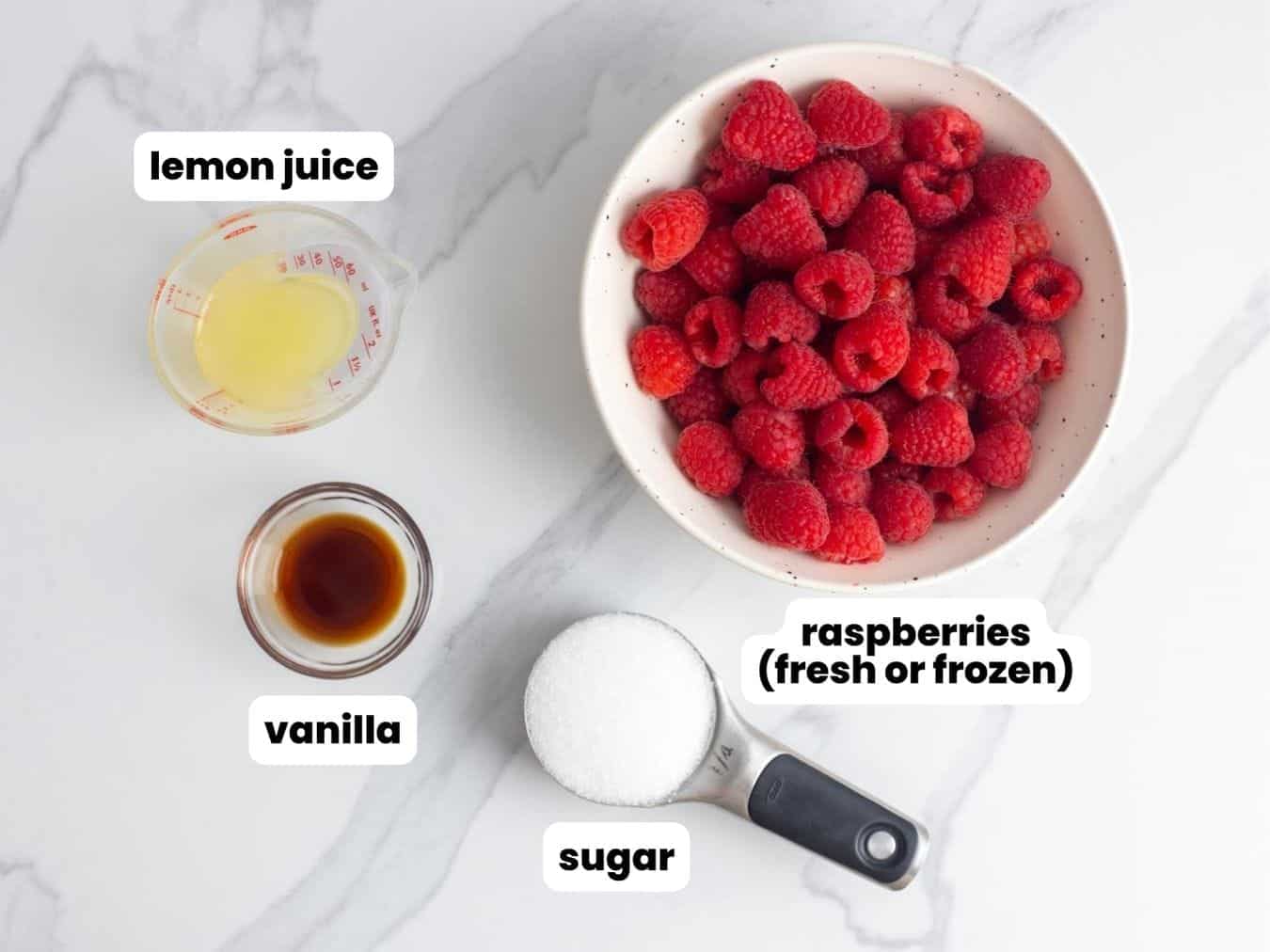 a bowl of fresh raspberries, a cup of sugar, a small bowl of vanilla extract, and a measuring cup of lemon juice, arranged on a marble counter.