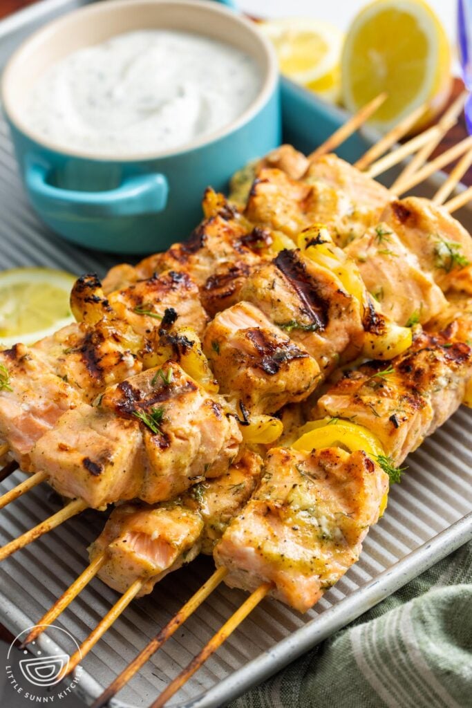 a small metal pan holding four salmon kabobs and a bowl of creamy salmon sauce.