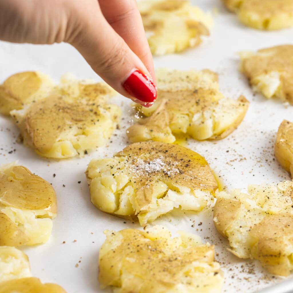 a hand with red fingernails adding salt and pepper to smashed potatoes on a tray.