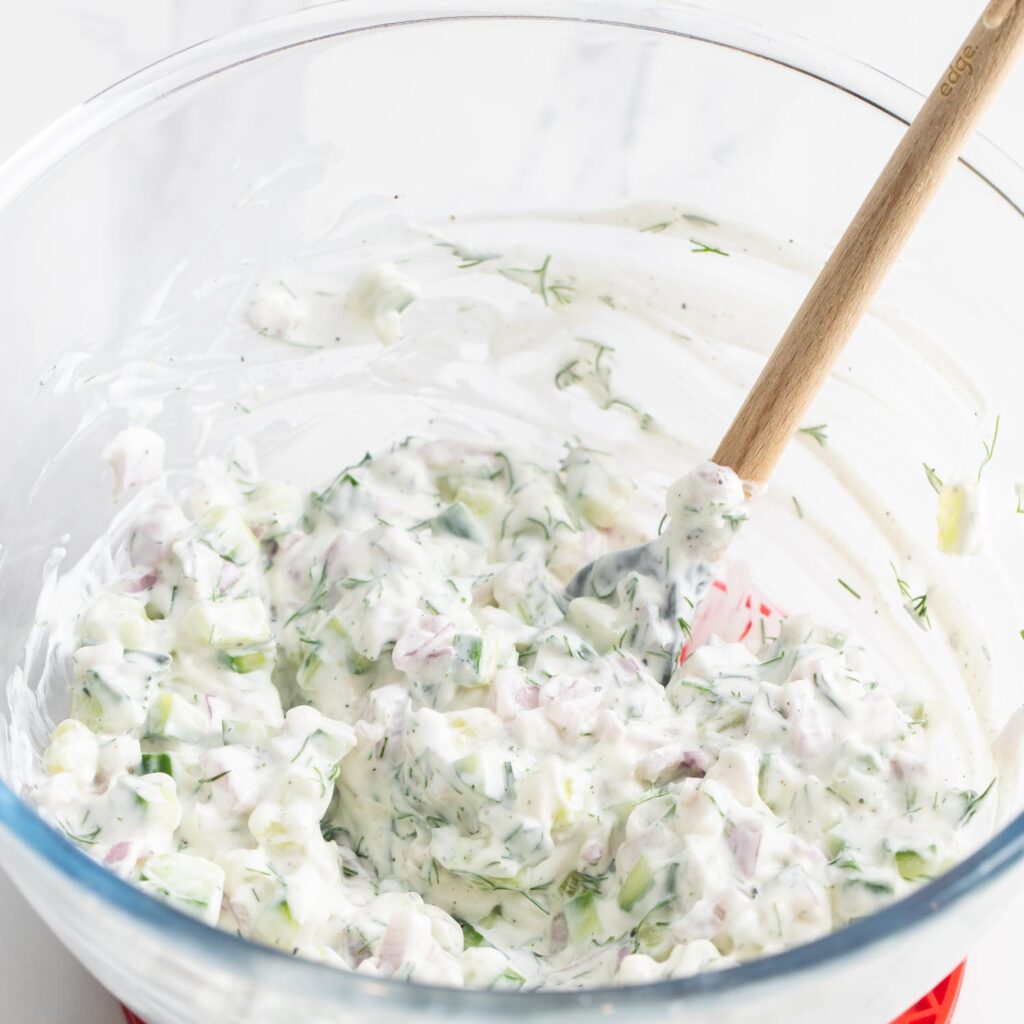 cucumber tzatziki sauce with dill stirred in a glass mixing bowl.