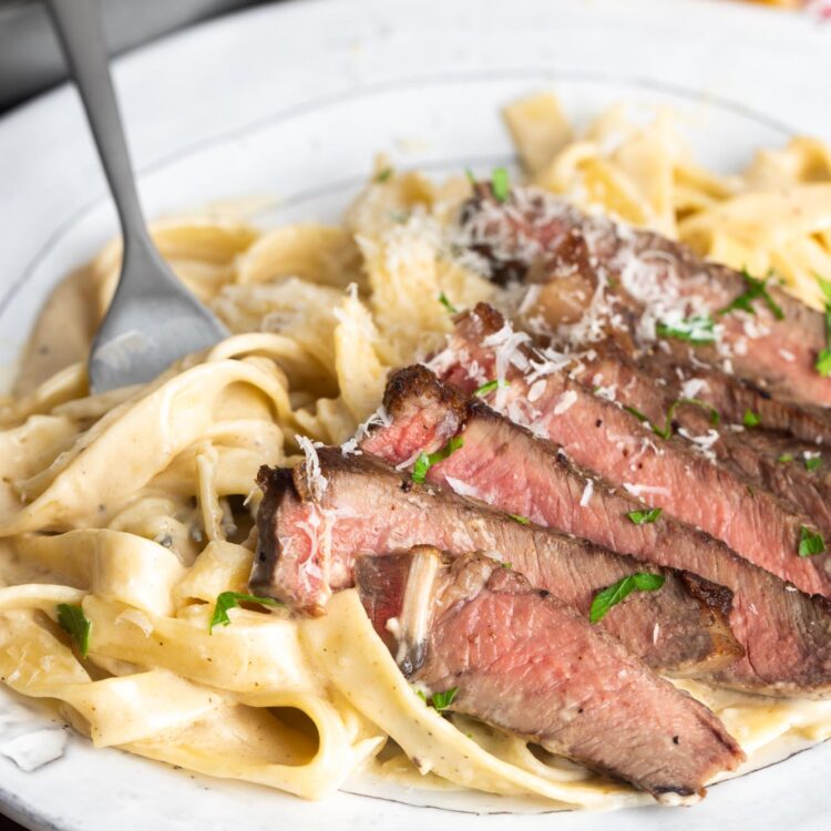 a plate of sliced steak with creamy pasta. A fork is picking up a bite of pasta.