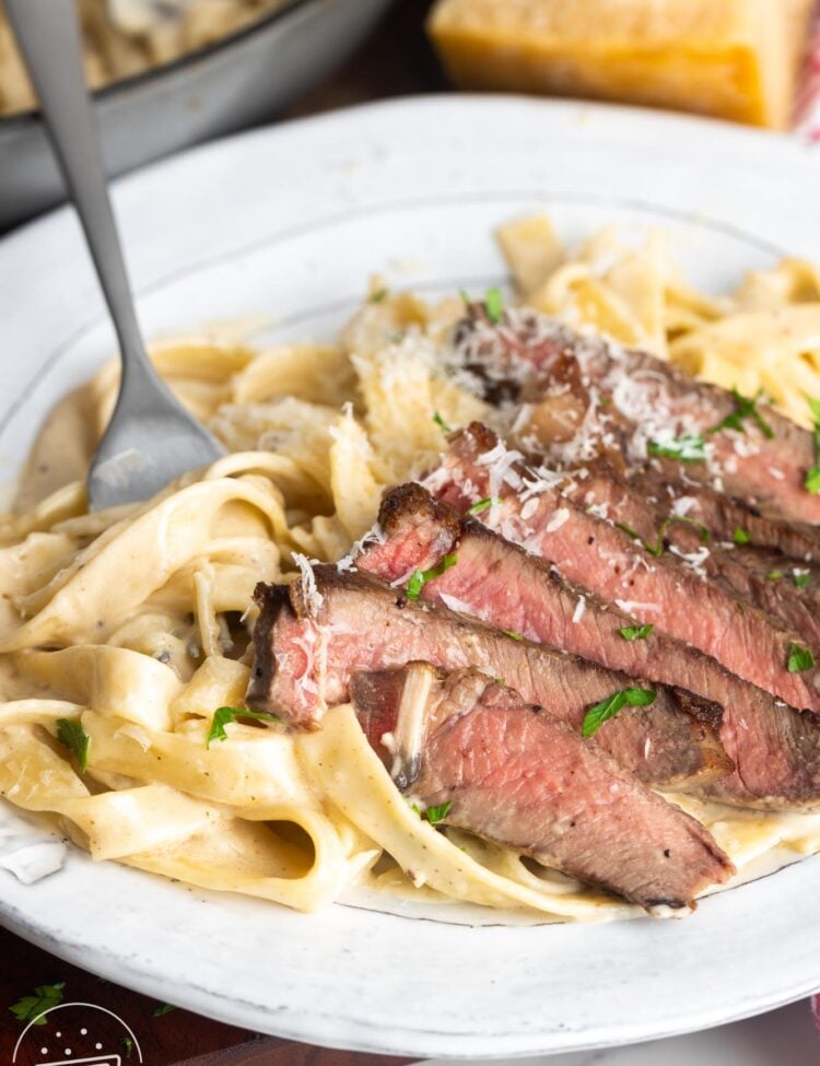 a plate of sliced steak with creamy pasta. A fork is picking up a bite of pasta.