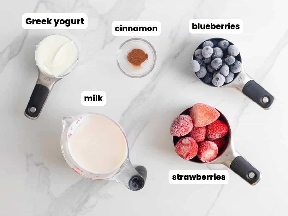 frozen strawberries, frozen blueberries, cinnamon, milk, and greek yogurt, all in separate measuring cups, on a marble counter.