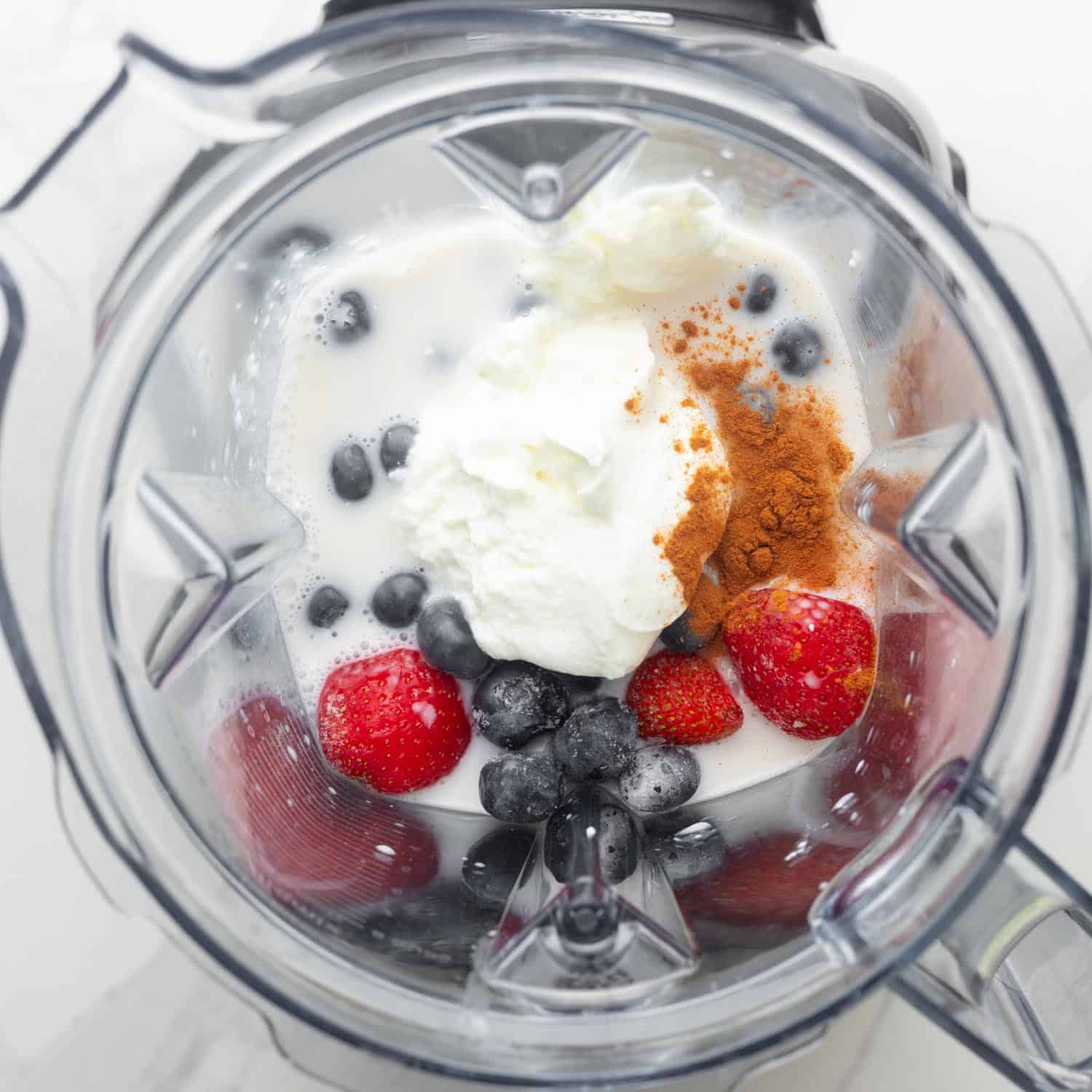 Ingredients for a strawberry blueberry smoothie with cinnamon and yogurt in a blender, viewed from above.